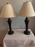 Pair of matching bedside lamps with dark bases and cream tapered fabric lampshades shown side by side on a surface against a plain wall.