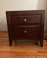 Frontal view of one wooden nightstand showing two drawers with metal knobs and curved fronts, dark brown finish.