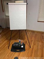 Full view of flip chart easel with a large white paper pad hanging, black tripod legs, and a black floor pad on the wooden floor. Items placed on pad include markers and spray bottle.