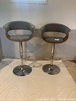 Two grey fabric and wood bar stools with round chrome pedestal bases and footrests shown side by side.