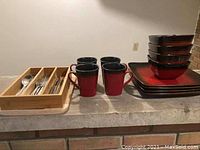 Photo showing four red and black mugs, bamboo drawer organizer filled with assorted metal cutlery and knives, and square dishes including bowls and plates stacked together.