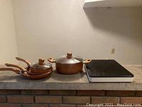 Photo showing the induction cooktop on the right and the set of two frying pans and two pots with lids on the left on a countertop.