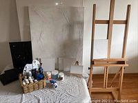 Full view of large wooden floor easel next to table with canvases and painting supplies including basket with paints and brushes