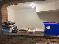 Wide view showing stacks of binders, large hole punch, file folders, paper stacks, and blue plastic file box with lid on brick surface under archway.