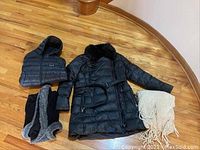 Full view of all items on floor showing black belted winter coat with fur collar, black hooded puffer jacket, black shawl, and cream knitted poncho shawl.