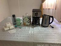 Full set of coffee and tea items displayed on table including coffee maker, kettle, mug, jar of teas, measuring spoons, and tea bags.