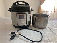 Full view of Instant Pot Duo Plus stainless steel and black plastic appliance with digital control panel, one inner cooking pot beside it, and power cord in front.
