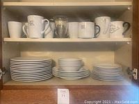 Shelved dishes organized in stacks showing plates, bowls, and mugs in two design styles (ribbed and plain). Mugs are assorted with different designs including text and geometric shapes.