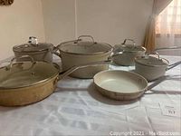 Overall view of 11-piece pot and pan set including various sizes of pots, saucepans, frying pans, and glass lids arranged on white surface.