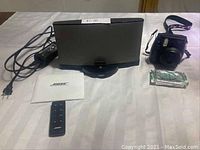 Wide shot showing Bose stereo speaker dock with iPhone charging connector, remote, power cable, Fujifilm Instax Mini 8 camera with wrist strap, and extra film pack on white textile surface.