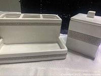 Photo of white ceramic bathroom counter items with rhinestone bands including toothbrush holder and covered canister, black wicker basket in background