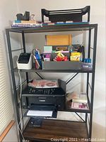 Full view of black metal frame shelving unit with various office supplies, Canon printer, label printer, books, and stationery on shelves.