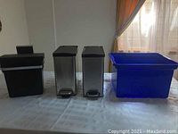 View of 2 stainless steel metal step cans with black lids and foot pedals next to a large blue recycling bin on a table.