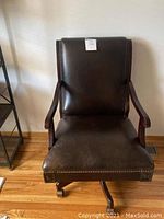 Front view of dark brown leather office chair with nailhead trim and wooden arms on castor wheels, showing wear and scratches on leather.