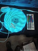 LED strip light powered on emitting blue light, showing remote control with multiple buttons for color and effect selection.