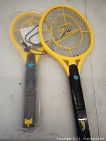 Two yellow electric mosquito swatters on grey surface, one outside and one in plastic packaging with instruction sheet and USB cable visible.