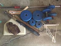 Photo of weights including multiple blue weight plates, dumbbells, two wooden baseball bats, Cooper Superblade boxed bat, Spalding baseball glove, and bike rack parts on concrete floor.