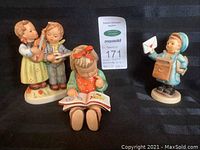 Three Hummel figurines displayed: "Happy Days" (boy and girl playing instruments), "Book Worm" (seated girl reading), and "Postman" (child in postal uniform holding a letter) shown together.