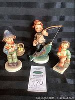 Three porcelain figurines displayed on black background. Includes Hummel Easter Greeting, Hummel Merry Wanderer, and Friedel Boy with Fish. Auction tag also visible.
