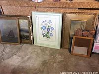 Wide view of six framed artwork pieces arranged on the floor against a wall, showing various subjects including sailing ships and floral painting.