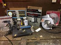 Lot overview photo showing multiple boxed and loose power tools including circular saw, scroll saws in boxes, heat gun box, impact driver kit, cans of paint remover on workbench