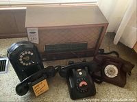 Four vintage items on carpet: one Philips wooden radio and three telephones including a black rotary wall phone and two tabletop rotary phones, one black and one brown.