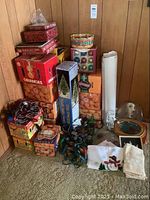Photo of stacked boxes of vintage Christmas decorations including lights, baubles, fiber optics tree, hand towels, and wrapping paper rolls on carpet floor in corner of room.
