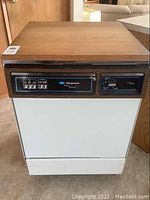 Front view of Hotpoint dishwasher showing wood panel top, white body, black control panel with buttons and knobs.