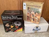 Photo showing boxed Charlescraft stainless steel electric steamer/skillet, boxed Revere Ware pots and pans, and boxed glass canisters with wood lids grouped together.
