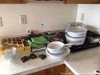 Overview of lot showing Corning Ware dishes, baking pans, cutlery, and kitchen items arranged on counter.