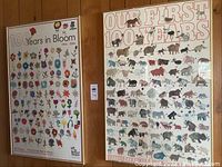 Photo shows two framed posters hanging on a wood-paneled wall: "100 Years in Bloom" with colorful flowers on left; "Our First 100 Years" with animal illustrations on right.