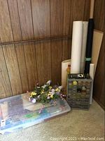 Wide angle photo of plastic bin with assorted fabrics, artificial flowers on top, gray storage unit with small drawers containing many thread spools, and large rolled fabrics against wood paneled wall.