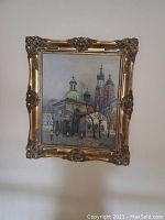 Full image of an ornate framed oil painting depicting a church with domed and spired towers, set among bare trees.