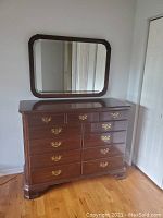 Wooden dresser with multiple drawers and brass handles, paired with a matching wall mirror with a dark brown wooden frame and rounded corners.