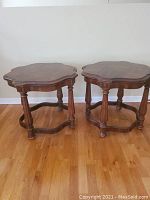 Pair of wooden end tables with scalloped edges and carved legs on hardwood floor against a white wall.