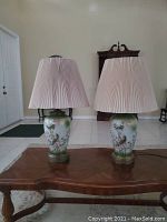 Pair of ceramic base table lamps with floral and bird design, brass accents, and pleated fabric shades on wooden table.