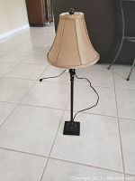 Full view of the 27-inch table lamp showing the beige fabric shade, dark metal stand, and cord lying coiled on a light tile floor.