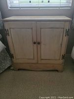 Photo showing the front view of a small white stained wooden cabinet with two paneled doors. The cabinet has metal handles and hinges on the sides and wooden knobs in the center. The cabinet appears slightly distressed with a rustic finish.