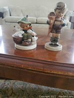 Two Hummel porcelain figurines placed on a wooden table with a sofa in the background.