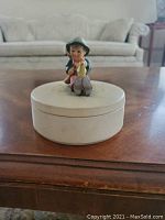 Front view of the round cream-colored candy dish with hinged lid topped by a figurine of a child in traditional Hummel style.