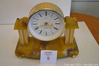 Front angled view of Art Deco mantle clock showing yellow marble look base, two side pillars, and round clock face with gold metal casing.