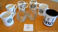Eight assorted mugs displayed on a wooden table, including Starbucks branded mugs, colorful sugar skull design mugs, clear etched glass mugs, and mugs with silhouette designs.
