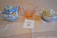 Photo shows three sugar bowls on a flat surface. From left, a blue-lidded floral porcelain sugar bowl with handle, a pink Depression glass sugar bowl with two handles, and a white porcelain floral sugar bowl with lid and dual handles.