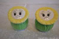 Two closed ceramic condiment jars with sunflower face lids, one smiling and one frowning, on a light patterned tablecloth.