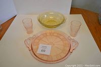 Overview of the lot showing pink cake plate with handles, pair of pink drinking glasses, and yellow Depression glass bowl on wooden surface