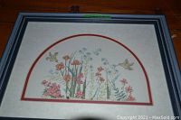 Close-up photo of the framed cross stitch garden scene with flowers and hummingbirds, showing detailed stitching and colors.