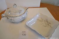 Full view of covered casserole with lid displaying black and grey leaf and fern decoration on white background with platinum accented handles and scalloped rim with plated edge. Next to square serving dish showing similar leaf decoration and scalloped edges.