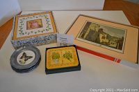 Four pewter coasters with butterfly designs, a boxed set of ceramic hot pads with botanical illustrations, a framed artwork and a boxed set of Jason herb coasters on a table.