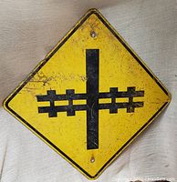 Front view of the vintage diamond-shaped railroad crossing sign, yellow with black graphic symbol of rail tracks crossing a vertical line, showing wear and rust.
