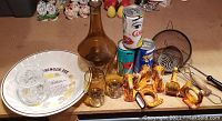 Overview of lot items on tabletop including collectible Coca-Cola cans, amber decanter set, ceramic napkin rings, porcelain plate, crystal glasses, and wire strainer basket.